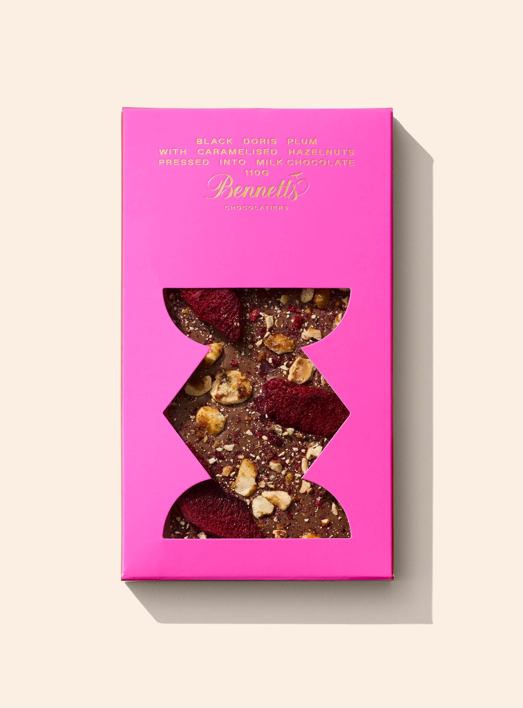Milk chocolate bar topped with caramelised hazelnuts and black doris plum pieces visible through a transparent window. Packaged in a magenta sleeve.