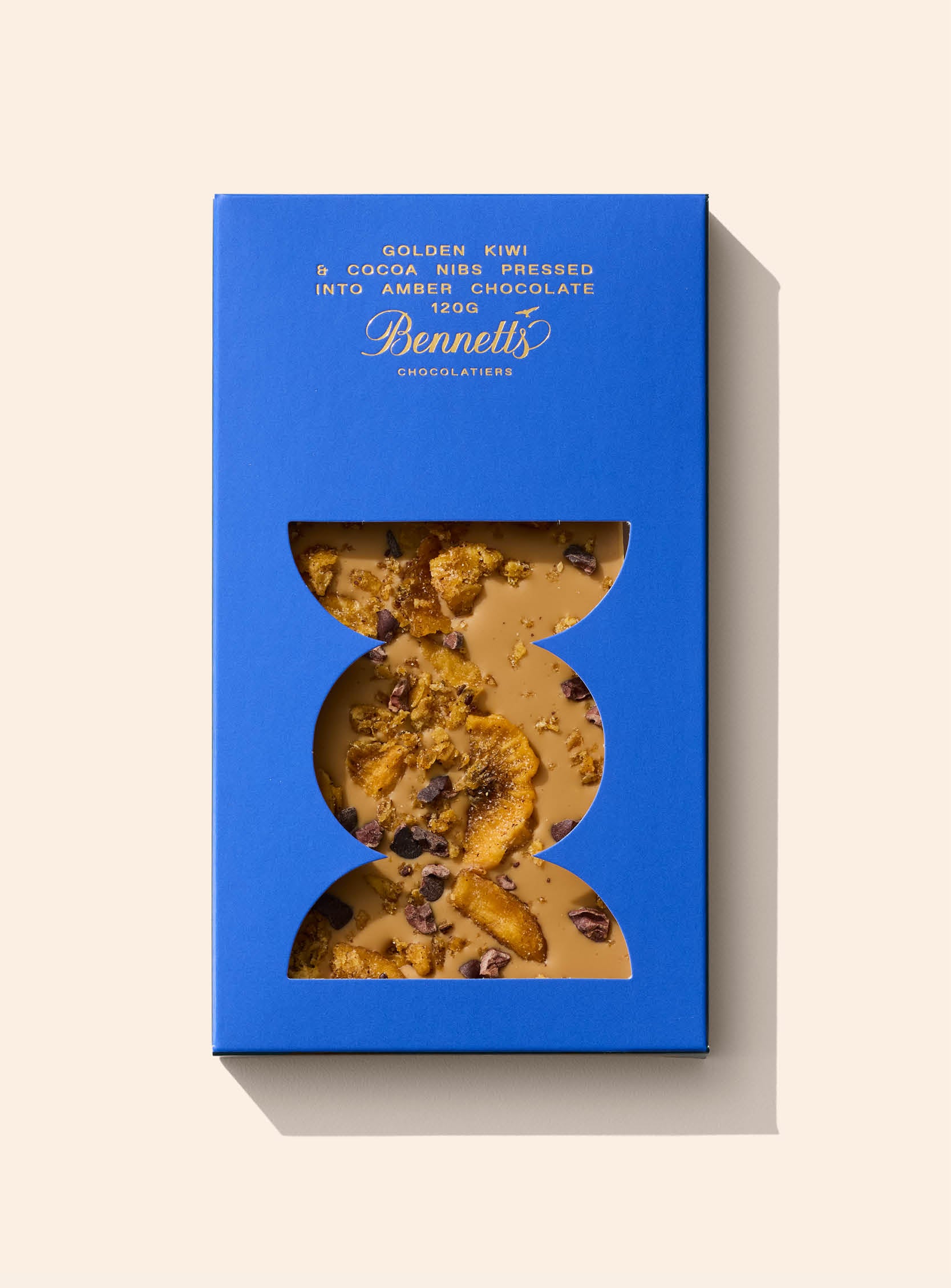 An amber chocolate bar with visible cacao nibs and kiwifruit pieces, packaged in a blue sleeve with a transparent window.